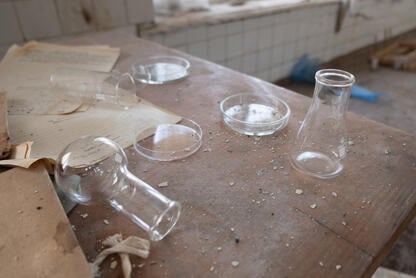 A photo of a dusty wooden table in an unused science laboratory; glass flasks, one knocked on its side, Petri dishes, and documents are strewn about the table. 