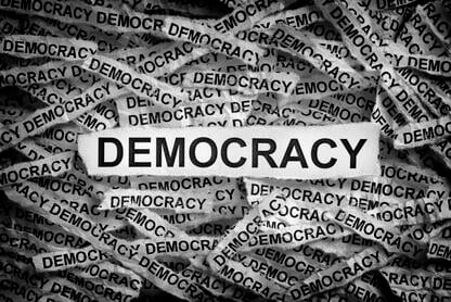 A black and white collage of torn-up strips of paper, each printed with the word "DEMOCRACY."
