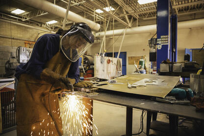 A young female student practices her welding skills in a technical classroom. She wears a face mask and sparks from the welding can be seen around her. 