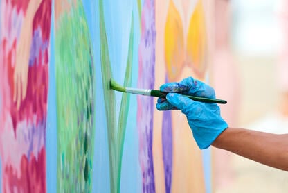 A photo of an artist's gloved hand holding a paintbrush and painting a brightly colored picture.