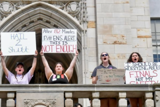 Four Pitt students stand on a balcony holding placards protesting the administration's slow response in notifying them of a reported active shooter threat.