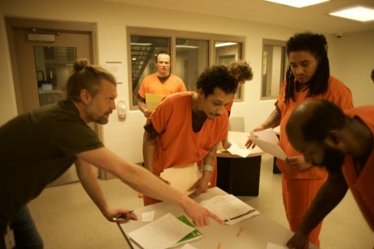 Nate Johnson, founder of FreeWriters, and three incarcerated students in orange uniforms stand around a table covered in papers.