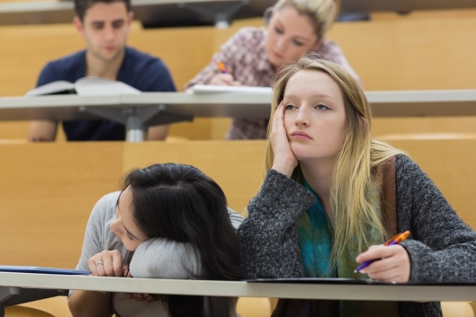 A group of disengaged, bored-looking students—one resting her head on her hand, and another with their head on the desk—in a university lecture hall.