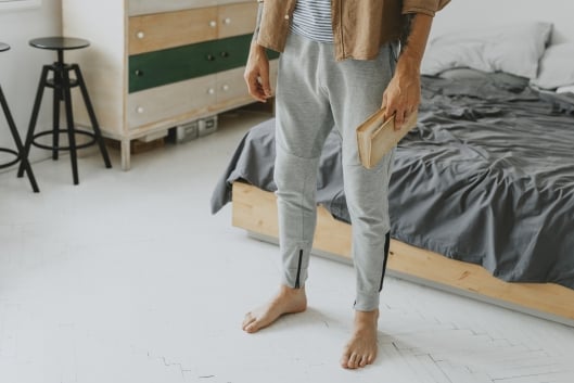 Student in bedroom with messy bed holding a book and wearing sweatpants 