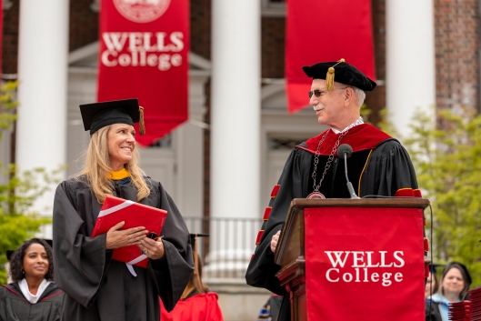 Wells College president Jonathan Gibralter, a light-skinned man with gray hair and a mustache, wearing academic regalia and sunglasses, on graduation day in front of red Wells College signage.