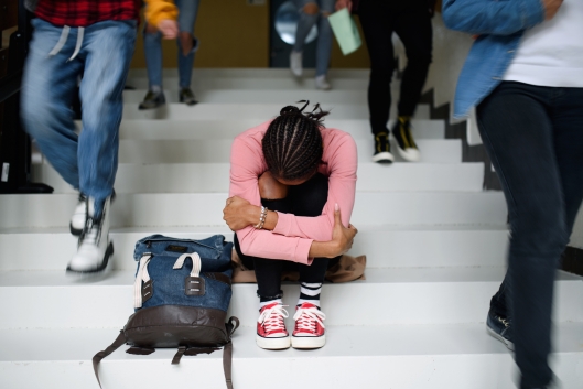 A student with her dark hair in braids sits on a staircase, hugging her arms around herself, as other students walk past