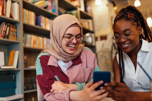 A Muslim student in a pink hijab shows her phone to another student, both smiling.