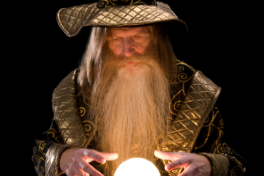 A wizard, dressed in a robe and pointy hat, stands with his hands hovering above a glowing crystal ball.
