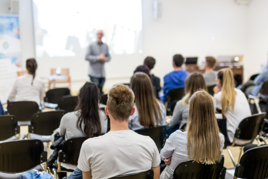 Students sit in a lecture hall facing the instructor at the front of the room