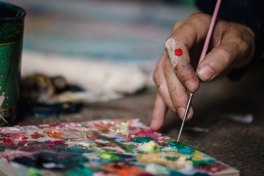 A hand with a paintbrush works on a colorful abstract painting on a wooden table.