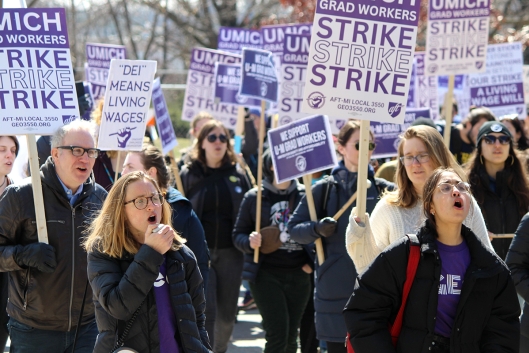 A photograph of striking University of Michigan graduate student workers, holding purple and white strike signs. 