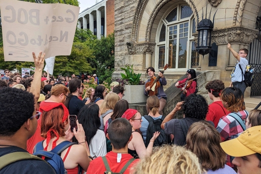 West Virginia University students protesting proposed faculty member layoffs and academic program cuts. One student holds a stick above the crowd, and another has a sign reading "Gee + BOG Need to GO."