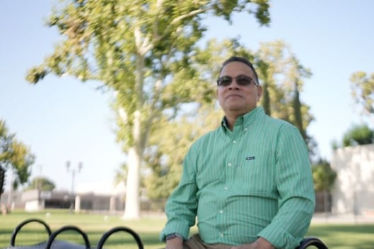 Daymon Johnson, a light-skinned man with dark hair wearing sunglasses and a long-sleeved green button-down shirt, sits on a bench looking out into the distance with trees behind him.