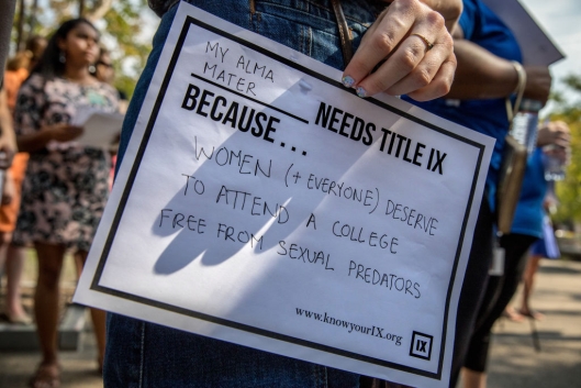 A person holds a sign on white paper that says "My alma mater needs Title IX because women and everyone deserve to attend a college free from sexual predators."