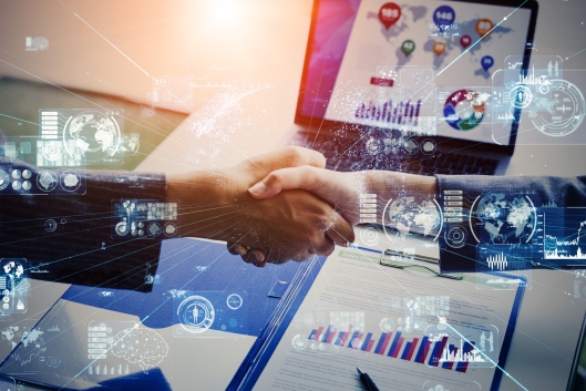Two male hands shake while there are blue "digital" icons floating around. A computer screen and paper bar chart are in the background.