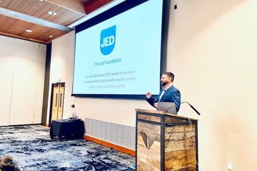 A man with a beard stands at a podium in front of a PowerPoint with the JED logo on it.