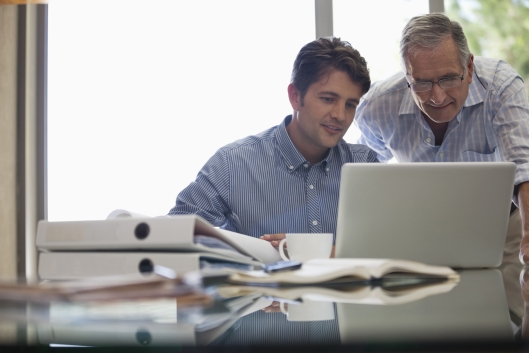 Older man leans over younger student who is looking at a computer and seems to be giving him advice