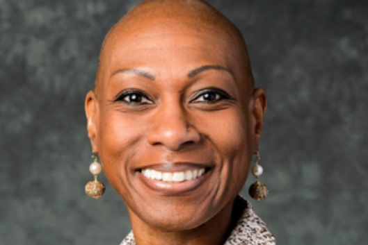 Antoinette "Bonnie" Candia-Bailey, a Black woman with a bald head, wearing a textured jacket and a statement necklace with matching earrings, smiles in a professional headshot.