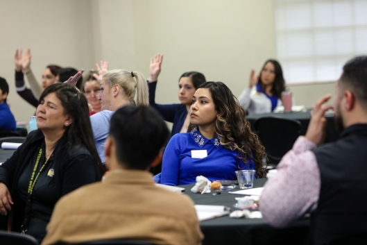 Faculty and staff sit at round tables, some raising their hands in the background. 