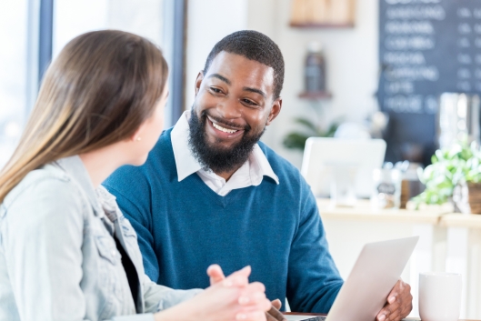 An African American man sits in a coffee shop with a woman and uses his laptop during their discussion.