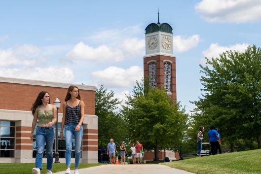 Two girls walk on a sidewalk with a college campus behind them. 