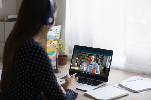 A woman wearing headphones faces a laptop. A man is on the screen of the laptop teaching. 