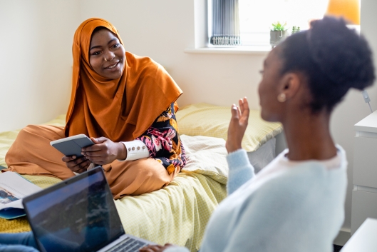 Two female university student friends in their dorm bedroom are studying together while sitting on the bed