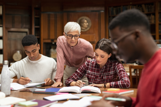 A group of three students studying at a table with the help of a professor standing behind them.