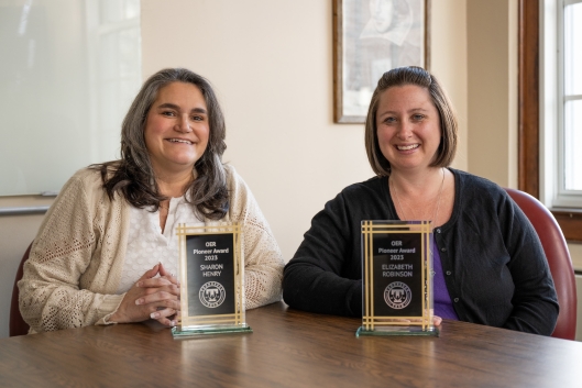Two female professors sit behind a desk with small awards on the desk. 