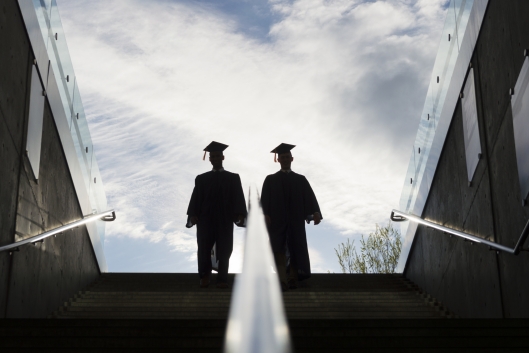 Two college graduates walk up a set of stairs, backlit with bright morning light.