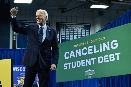 President stands in a suit jacket and tie holding a thumb up. A sign saying, “Canceling Student Debt,” is displayed behind him.
