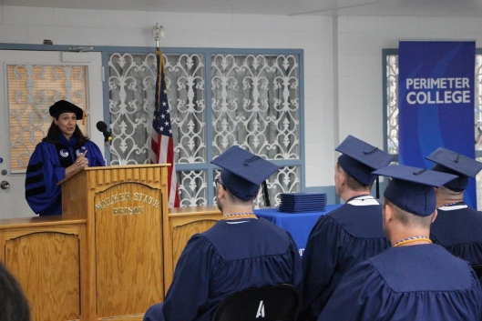 A group of students in caps and gowns sits in front of a professor at a podium. A blue “Perimeter College” banner is in the background.
