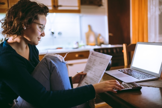 A student sits at her kitchen table with bills and a calculator, laptop open in front of her