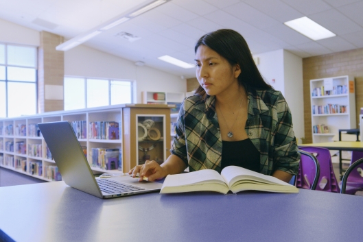 A student in a library works on her laptop with a book open next to her