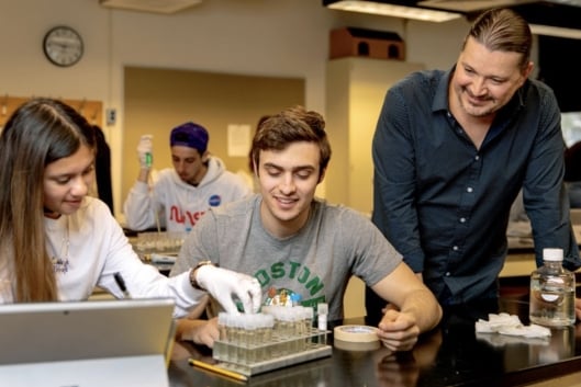 Mark Sarvary stands next to two seated students working with test tubes in a lab course setting, smiling. 