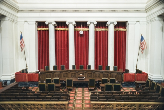 A photo of the interior of the U.S. Supreme Court chambers, showing a curved table with nine chairs in front of a set of five long red curtains hanging between four white columns.
