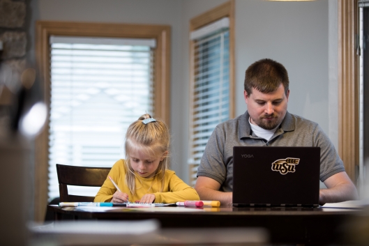 An adult student works at a laptop while his young daughter colors next to him