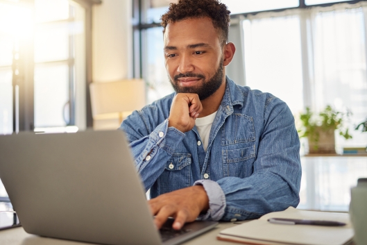 An adult man works at a laptop with a notebook and pen beside him.