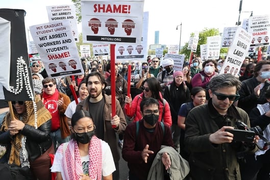 A photograph of a crowd of protesters, some holding signs bearing the name of the Massachusetts Institute of Technology Graduate Student Union.