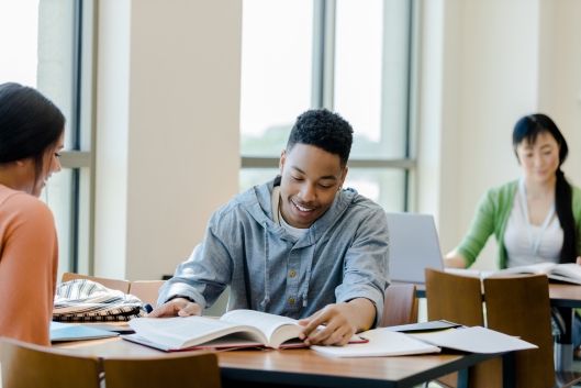 The young adult male receives tutoring in the library from a peer so he can better understand the material.