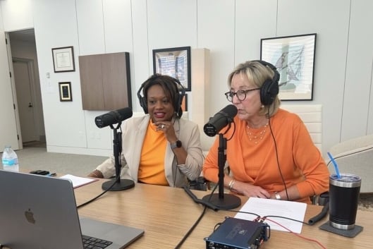 Amber Williams and Donde Plowman sit next to each other, wearing University of Tennessee orange, with microphones at the table in preparation for a podcast interview.
