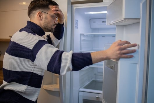 A young man holds a fridge door open and leans against the fridge, his head in his hand, as he surveys its contents: the fridge is empty. 