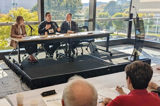 A photograph of three people on a panel sitting at a table, with large glass windows behind them.