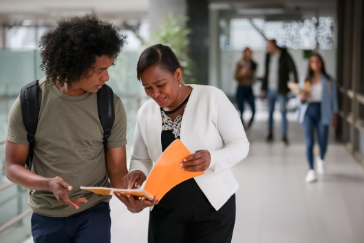 Happy teacher talking to an African American college student in the hall at the school and pointing at his notebook