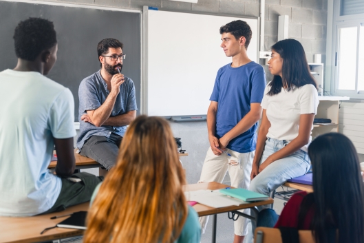 A male instructor engages in discussion with a small group of five students variously standing/sitting in a circle.