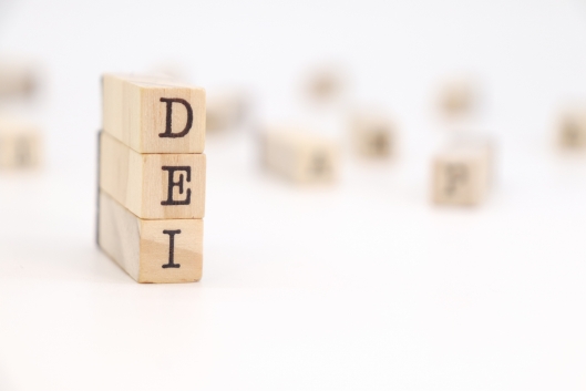A vertical stack of three wooden blocks with letters on them reads "DEI." Other blocks, their letters blurry, can be seen strewed in the background.