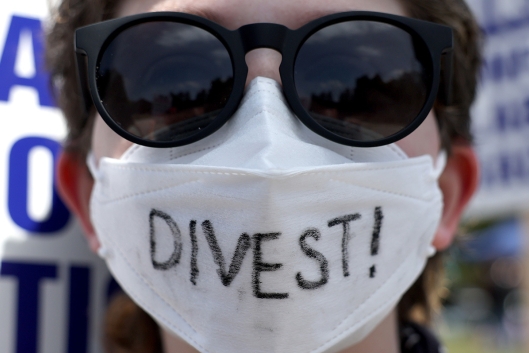 A photograph of the face of a person wearing sunglasses and a white face mask, on which "Divest!" is written. 