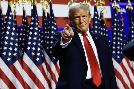 Donald Trump, in a suit and red tie, stands in front of a row of American flags and points his finger.