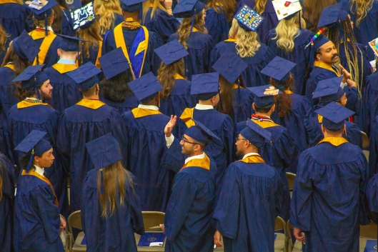 A group of students in blue and yellow graduation regalia face away from the camera. One student turns to the left to wave to someone out of frame.