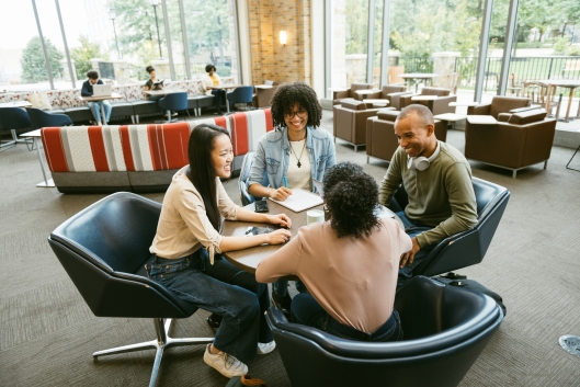 In a student center lounge space, four students sit around a table as if running a meeting. 